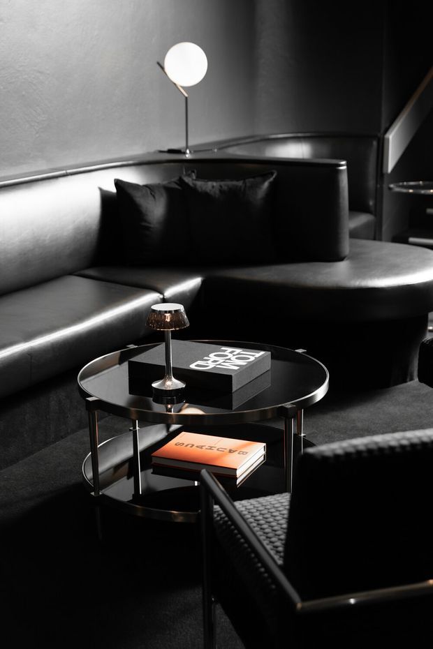 A dramatic, monochromatic living room or lounge with a large black leather sectional sofa, matching dark cushions, a round black glass coffee table, sculptural table lamp, books, and a textured armchair in the foreground. The space has dark walls, reflective surfaces, and low, focused lighting, creating a refined, cinematic atmosphere.