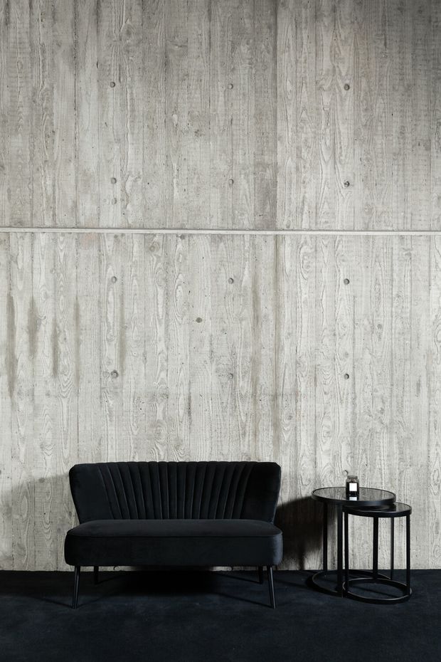 A minimalist interior vignette featuring a black upholstered, channel-tufted sofa and two small round black metal side tables against a pale grey vertically textured timber wall. The dark floor and monochromatic palette create a moody, refined atmosphere with strong contrast.