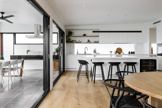 Bright contemporary open-plan kitchen and dining area with white flat-panel cabinetry, a long island with four black bar stools, integrated appliances, light timber flooring, and a round timber dining table. Black-framed glazed sliding doors open to a tiled dining or living area with additional white cabinetry and bar seating. The room has a clean, airy palette with subtle greenery and black accents.