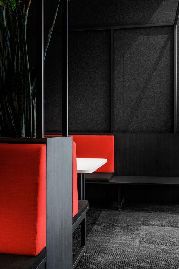A dramatic, compact commercial interior featuring red upholstered booth seating, black built-in banquette seating, dark charcoal acoustic wall panels with black framing, a small white table, and dark patterned carpet. Tall indoor plants appear at the edge of the space. The palette is predominantly black, charcoal, red, and white, creating a bold contemporary hospitality or dining setting.