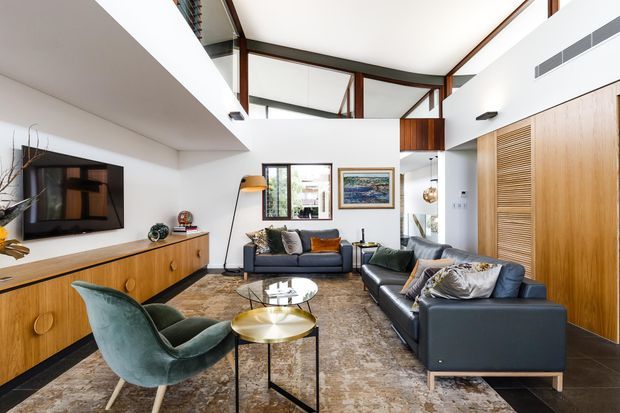 A bright, open-plan contemporary living room with a double-height ceiling and clerestory glazing. The room features white walls, exposed dark timber ceiling beams, extensive timber built-in cabinetry and louvred doors, dark leather sofas, a green upholstered lounge chair, and layered round coffee tables over a large patterned rug. A television and low timber media console line the left wall, with artwork and glazing at the rear.