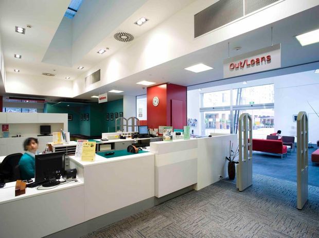 Bright modern bank or financial services branch interior with a white reception and customer service counter in the foreground, computer workstations, signage, security gates, and a waiting area with red seating beyond. The space has a clean commercial fit-out, white surfaces, grey carpet, red feature walls, extensive glazing, and a high ceiling with recessed lighting and ventilation grilles.