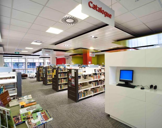 Bright modern library interior with rows of bookshelves, catalogue and section signage, a white built-in computer workstation, patterned carpet, colourful wall panels, suspended acoustic ceiling, and glazed entrance doors.