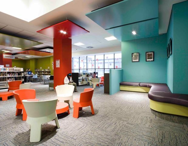 A colourful contemporary library or educational reading space with open shelving, study areas, lounge seating, bright orange and white chairs, teal walls, a red structural column, and a patterned carpet. The space is open and brightly lit, with built-in curved bench seating along the right wall and large windows at the rear.