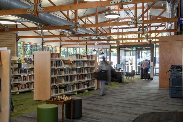 A contemporary library interior with rows of bookshelves, reading and study areas, a reception or service desk near glazed entry doors, and people moving through the space. The room has an exposed timber post-and-beam ceiling with metal bracing, visible silver ventilation ducts, suspended circular lighting, carpeted flooring, and large windows providing abundant daylight. A small chess table and stools occupy the foreground.