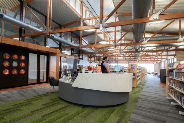 A bright, open-plan library or commercial learning space with a curved reception desk in the foreground, extensive bookshelves, exposed timber roof trusses, visible metal bracing and large HVAC ductwork. The floor combines grey carpet with green carpet bands, while glazed partitions and clerestory windows bring in natural light. A dark feature wall with circular displays and a small lounge chair sit to the left of the reception area.
