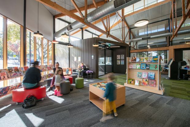 Bright, open-plan public library or community reading space with large windows, exposed timber roof trusses, corrugated metal ceiling, exposed ductwork, suspended pendant lighting, carpeted floors, colourful modular seating, low timber display shelving, and a children's book area. A darker enclosed room and additional shelving are visible toward the rear.