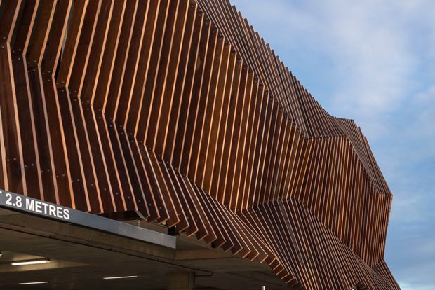 Close-up exterior view of a contemporary building facade formed by dense, repeating vertical timber-toned fins. The fins create a dramatic folded and faceted geometric surface above a concrete vehicle entry, with a visible 2.8-metre clearance sign and exposed soffit lighting. Blue sky is visible behind the structure.