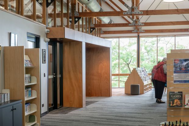 A bright, double-height library or community reading space with exposed timber post-and-beam structure, visible metal bracing, corrugated services ductwork, extensive glazing, and built-in timber shelving and cabinetry. The carpeted floor and open-plan layout create a welcoming public interior, with bookshelves, display stands, and a person browsing at the right.