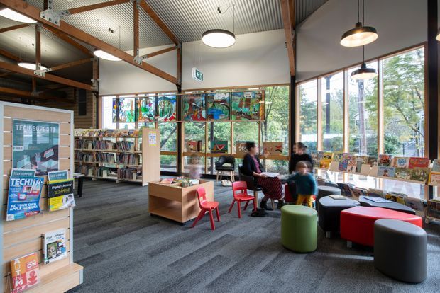 A bright, contemporary public library or community reading area with floor-to-ceiling windows, bookshelves, children's seating, display shelving, and colourful stained-glass or graphic window panels. The space has an exposed timber-and-steel structural ceiling, corrugated metal ceiling lining, suspended pendant lights, and dark carpet flooring. People are seated in the reading area, with trees visible outside.
