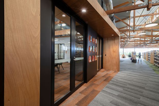 Interior view of a contemporary commercial library or learning space with exposed timber structure and services, dark-framed glazed office doors, warm timber wall and ceiling lining, and broad carpeted circulation areas. Bookshelves and workspaces are visible in the open area beyond.