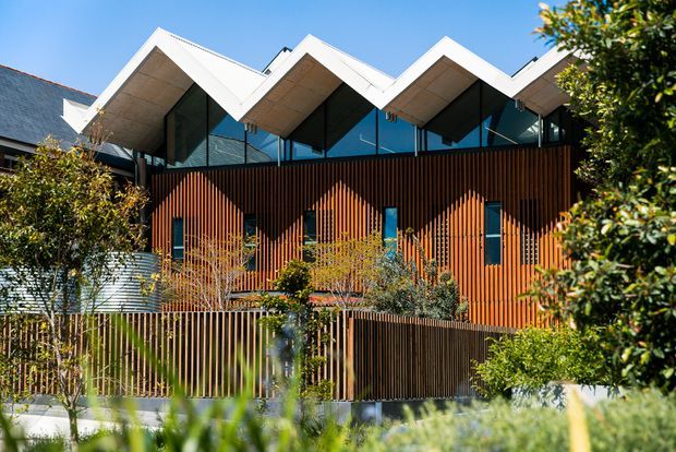 Exterior view of a contemporary multi-level building with a distinctive repeated zigzag roofline, deep white roof overhangs, extensive upper-level glazing, and a warm vertical timber-screen facade. Landscaping and a matching timber boundary fence sit in the foreground.