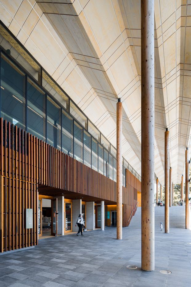 A covered commercial building forecourt or colonnaded entrance with tall timber columns, a pale tiled or panelled ceiling and soffit, extensive glazed upper-level windows, and a vertical timber batten facade. Broad grey stone paving leads past retail or institutional entrances and a stairway. The architecture is contemporary and civic in character.