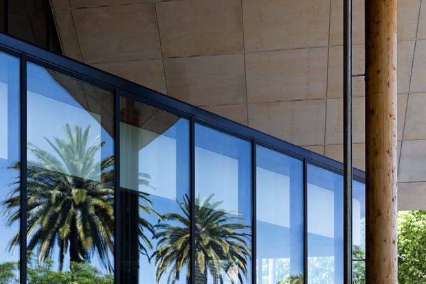 Close-up architectural view of a modern glazed facade beneath a broad soffit, with dark-framed floor-to-ceiling windows reflecting palm trees. A substantial round timber column supports the covered structure, and warm-toned panelled ceiling surfaces are visible.