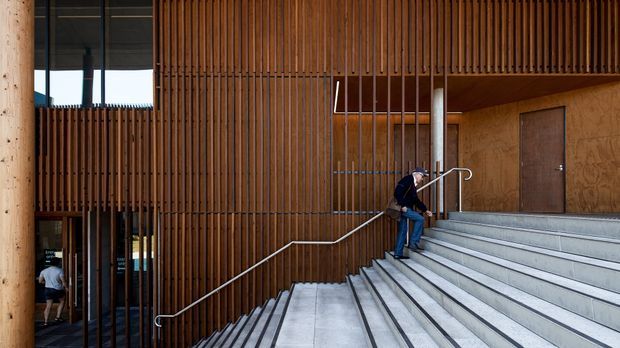 Exterior architectural view of a contemporary multi-level building entrance, featuring broad concrete steps, slim metal handrails, warm vertical timber screening, dark-framed glazing, and a sheltered upper-level walkway. A person is seated on the stairs, emphasizing the scale of the public-facing entry.