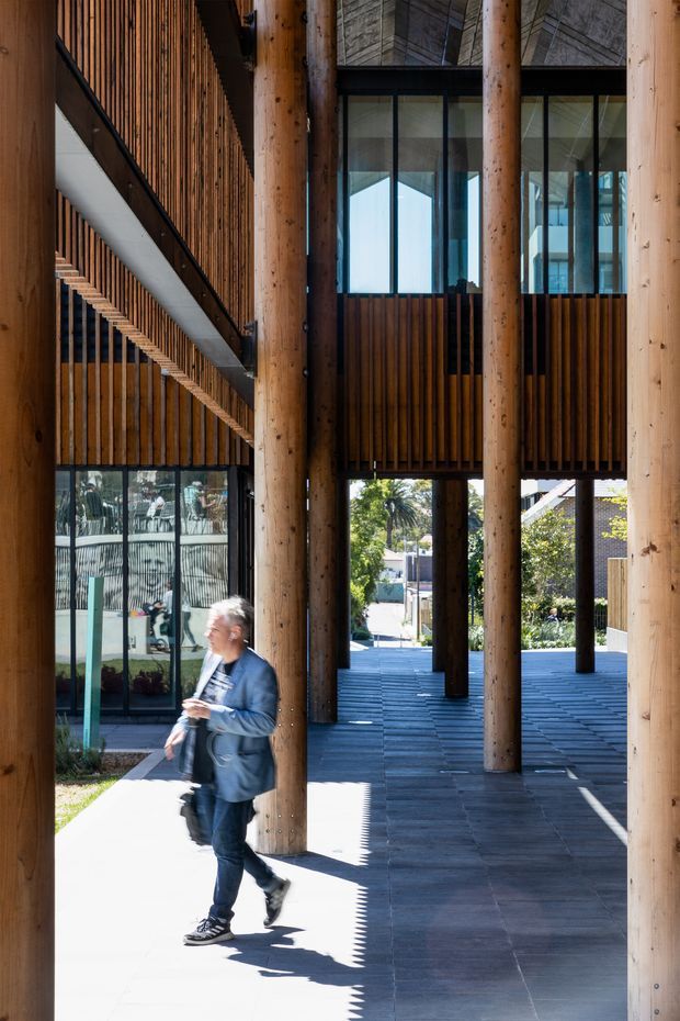 A tall, multi-level architectural atrium or covered walkway with prominent round timber columns, vertical timber slat screens, dark tiled paving, and extensive floor-to-ceiling glazing. A stair or ramp runs along the left side, while open passages connect to an outdoor landscaped area beyond. A person is walking through the space. The design combines exposed timber structure with a contemporary institutional or commercial character.