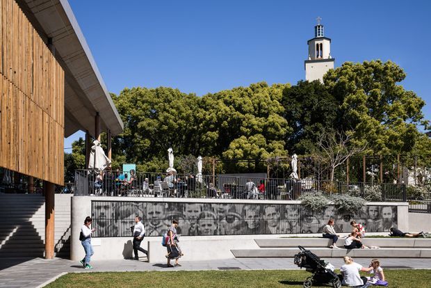 A contemporary public or commercial complex with a timber-clad building and broad concrete steps at left, overlooking an outdoor terrace with café seating, umbrellas, railings, and a large monochrome mural. People gather around stepped seating in a landscaped urban setting, with a distinctive tower visible above the trees in the background.