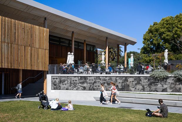 A contemporary low-rise pavilion-style building with a broad projecting roof, timber slat screening, dark glazing, slender structural columns, and a raised outdoor terrace. The terrace includes café-style seating and umbrellas, while a landscaped lawn and pedestrian forecourt occupy the foreground. A patterned mural or screened graphic appears along the terrace retaining wall.