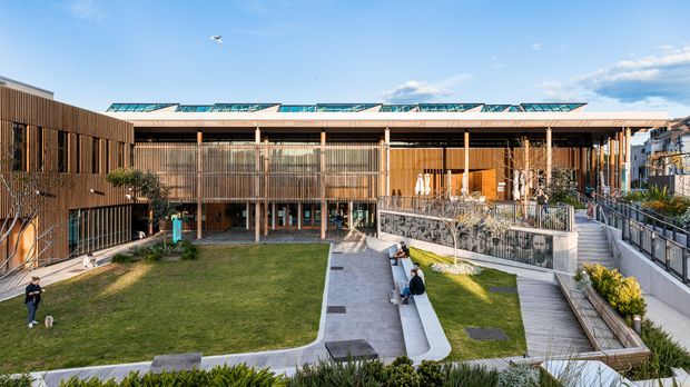 Wide exterior view of a contemporary multi-level building arranged around landscaped courtyards and pedestrian paths. The facade combines warm vertical timber cladding, extensive glazing, screened balconies, slender columns, and a low roof with visible rooftop glazing or skylight structures. Lawns, raised planters, timber decking, and concrete walkways occupy the foreground.