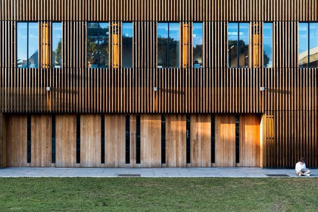 A two-storey contemporary building facade clad in vertical timber slats and broad timber panels. Tall narrow windows are integrated into the upper level, while the lower level has large timber doors or panels with slim dark openings. A paved path and lawn sit in front of the building, with a person seated at the far right for scale.