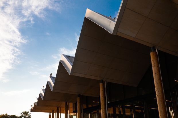 Low-angle exterior view of a contemporary building with a dramatic stepped, projecting roofline supported by timber posts. Dark glazing sits beneath the deep overhangs, with a bright blue sky and a few palm trees in the background.