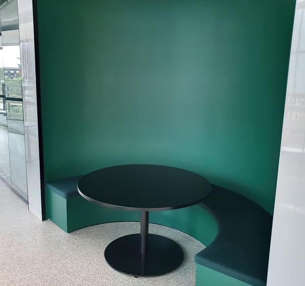A compact contemporary built-in seating nook with a curved dark green upholstered bench wrapping around a deep green feature wall. A small round black pedestal table sits in front, with glossy white and glass architectural surfaces visible at the sides. The space is finished with a speckled light terrazzo-like floor and a bold, refined colour-blocked palette.