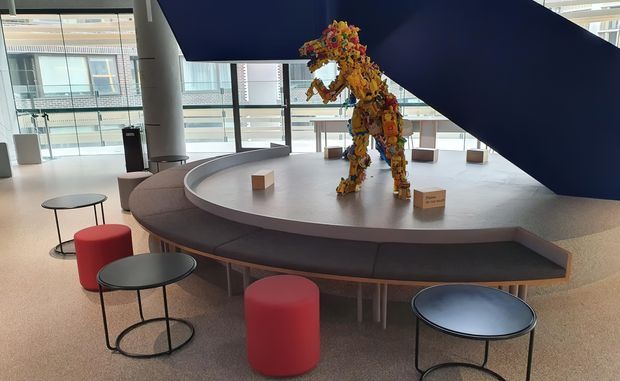 Bright contemporary commercial atrium or lobby with expansive glazing, a sculptural curved built-in seating platform, small round tables, red upholstered stools, and a colourful abstract animal sculpture beneath a large dark-blue geometric overhead feature.