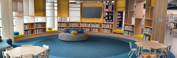 A bright contemporary library or reading area with curved built-in timber shelving, colourful yellow and blue wall panels, a large central display screen, blue carpet, low rounded seating, and small timber tables and chairs. The space appears designed for children or students, with bookshelves and flexible reading and study zones.