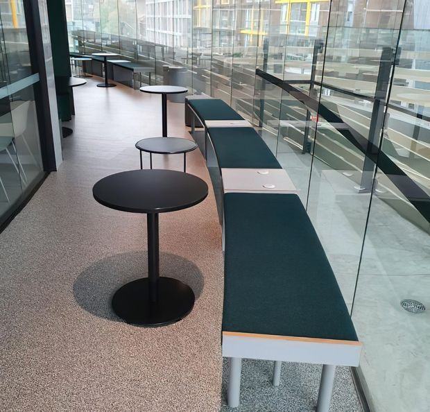 A contemporary commercial interior showing a long glazed corridor or informal seating area. A dark upholstered bench runs beside a full-height glass balustrade, accompanied by small round black café tables. The space has a terrazzo-like speckled floor, dark framing, and views into a multi-level atrium or adjacent building through the glazing.