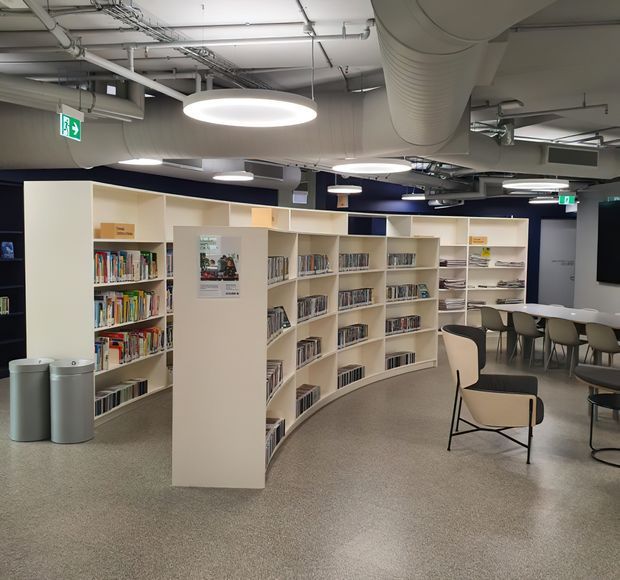 A contemporary library or learning commons with curved cream-coloured shelving filled with books, reading tables and chairs, and a large wall-mounted display. The space has an exposed services ceiling with ducts, pipes, and suspended circular pendant lights, polished speckled flooring, and dark accent walls.