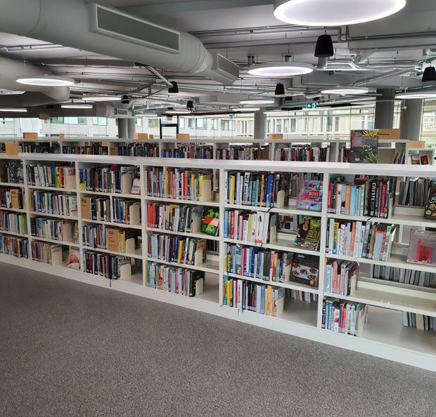 A bright open-plan library or educational resource space with long rows of white built-in bookshelves filled with books. The room has a grey carpeted floor, exposed ceiling services and ductwork, suspended circular pendant lights, structural columns, and large windows. The overall appearance is functional and contemporary with an industrial-style exposed ceiling.