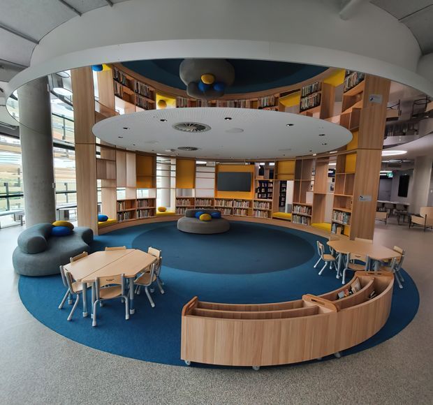 A vibrant modern library or reading area with curved timber shelving, integrated circular seating, blue carpet, small tables and chairs, and a suspended oval ceiling feature. The space has a playful, child-friendly character with colourful cushions and strong curved forms, plus large glazed openings and a mezzanine-like upper level visible at the edges.
