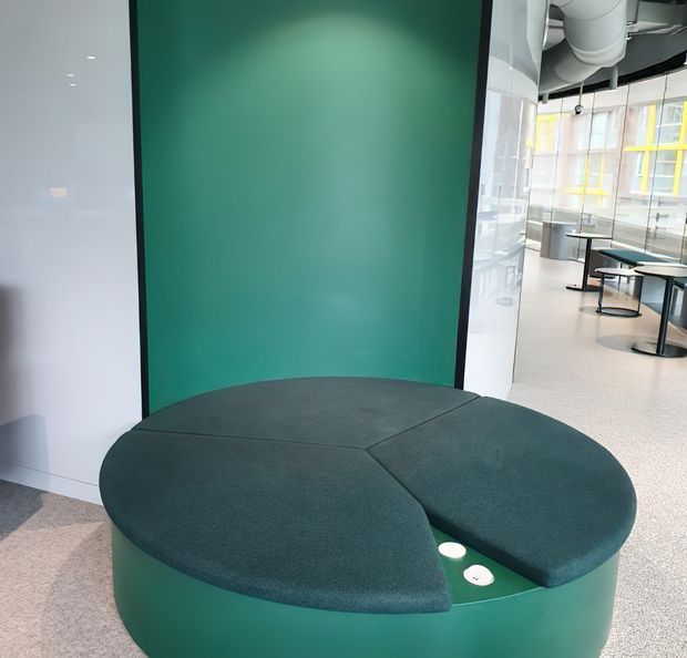 A contemporary commercial interior featuring a sculptural, rounded green upholstered seating pod in the foreground, set against a curved deep-green wall panel. Behind it is an open breakout or lounge area with small round tables, stools, glazed partitions, exposed ductwork, and terrazzo-like flooring.
