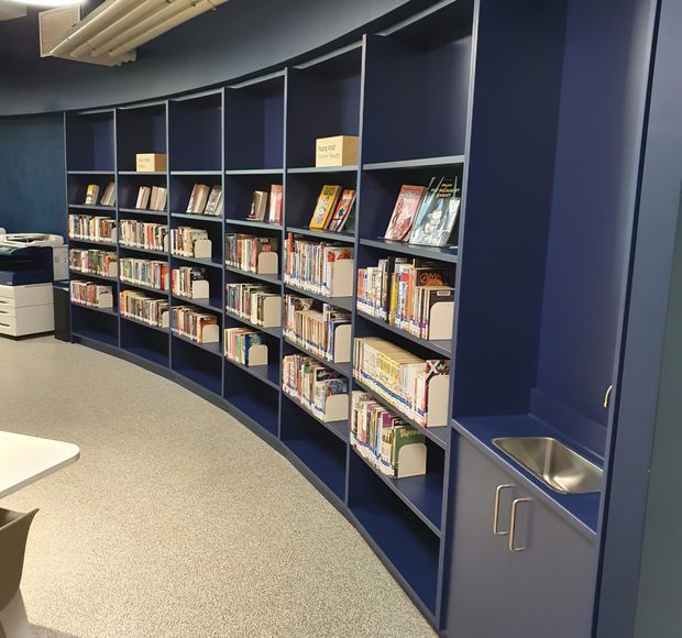 A curved wall of deep navy built-in library shelving filled with books, magazines, and display items. The space includes a small integrated stainless-steel sink and cabinetry at the right, a printer or copier at the left, a neutral speckled floor, and exposed linear ceiling services. The image appears to show a contemporary library or reading area.