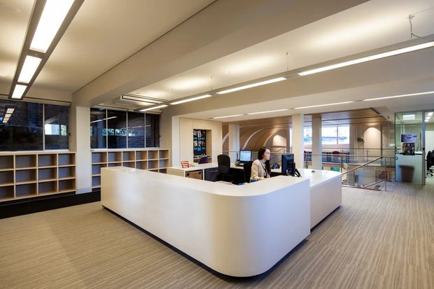 A bright contemporary reception area with a large white curved-front reception desk, computer workstations, open shelving, carpeted flooring, extensive glazing, and linear suspended and recessed lighting. The space opens toward a mezzanine or stairwell area and appears to be a professional office or commercial interior.