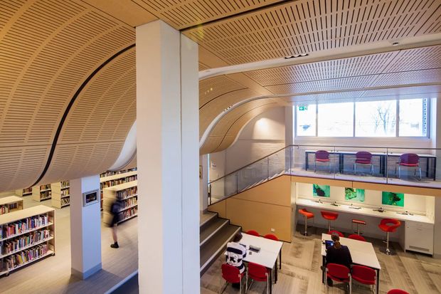 Interior view of a contemporary multi-level library or learning space with sweeping curved timber slatted ceilings and wall panels, white structural columns, bookshelves, a stairway, mezzanine-level seating, and collaborative tables with red chairs. The space is brightly lit by large windows and features integrated study benches and shelving.