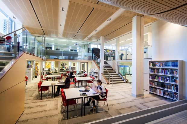 A bright, multi-level library or learning commons interior with rows of study tables and red chairs, bookshelves, staircases, glass balustrades, and a prominent slatted timber acoustic ceiling. The space has abundant daylight, white structural columns, carpeted flooring, and an open institutional layout.
