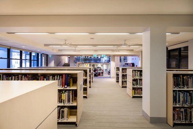 Wide interior view of a contemporary library or institutional reading space, with long rows of low and tall bookshelves, a broad carpeted circulation aisle, large glazed windows, white structural columns, suspended linear lighting, and ceiling fans. The space is brightly and evenly lit with a clean, functional fit-out.