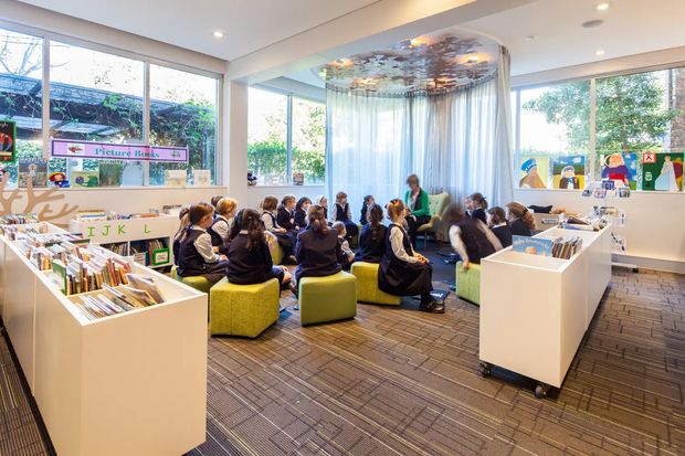 Bright contemporary school library or reading area with large windows, white built-in and freestanding book shelving, a group of children seated on colourful ottomans, and a teacher or librarian reading near a curtained circular nook. The room has patterned carpet flooring, white walls and ceiling, and a decorative ceiling feature above the reading area.