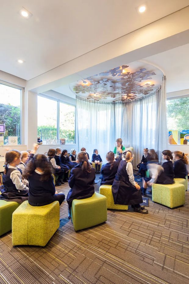 Bright contemporary educational or community interior with a group of children seated in a circular discussion area. The room features expansive windows with sheer curtains, a patterned decorative ceiling installation with integrated pendant lights, white walls and beams, green upholstered cube seating, and patterned carpet flooring.