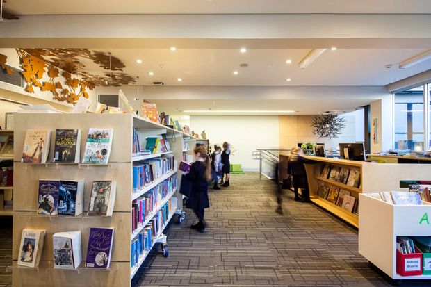 A bright contemporary library or bookstore-like commercial interior with open shelving displaying books, a central circulation or service desk, carpeted flooring, recessed ceiling lighting, and several people browsing or working. Large windows provide daylight, while light timber and white finishes create an open, welcoming public-space atmosphere.