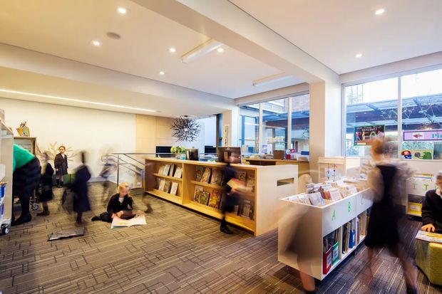 Bright contemporary commercial interior, likely a library or bookstore, with open shelving, display stands, books and magazines, large windows, recessed ceiling lighting, and people moving through the space. The room has a warm neutral palette and patterned carpet flooring.