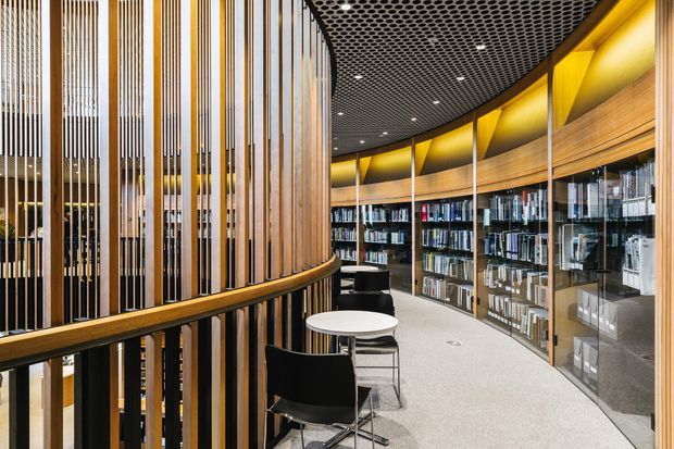 Curved contemporary library or reading area with extensive built-in bookshelves behind glass, small round tables and black chairs, a dramatic black perforated ceiling, warm timber wall panelling, and vertical timber screening around a curved balustrade. The space has a refined, warmly lit commercial or institutional interior character.