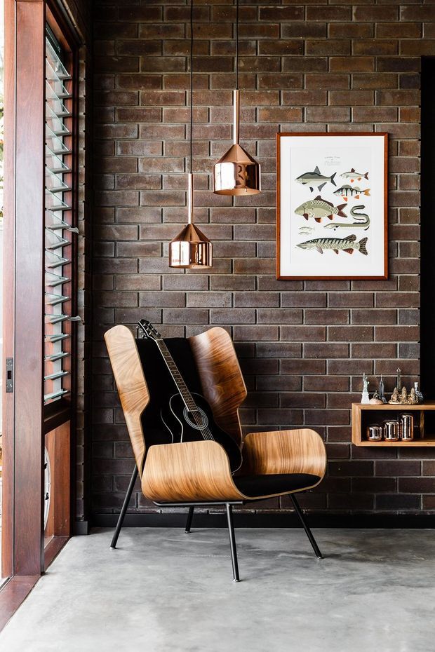 A stylish interior vignette featuring a mid-century-inspired wooden lounge chair with black upholstery and a black acoustic guitar, set against an exposed dark brick wall. Two copper pendant lights hang above, while a framed fish illustration and a small wall-mounted timber shelf with decorative objects add character. A louvred timber-framed window is visible on the left, and the floor is polished concrete.