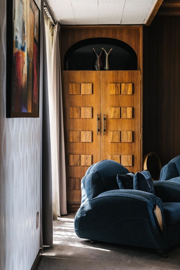 A richly styled interior corner featuring a tall, dark timber built-in cabinet with geometric drawer fronts and an arched display niche. Deep blue sculptural lounge chairs occupy the foreground, while patterned wallcovering, heavy curtains, framed artwork, and a carpeted floor create a dramatic, vintage-inspired sitting area.