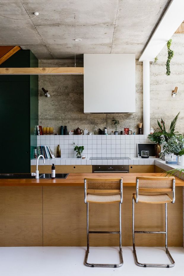 A compact contemporary kitchen with a timber island and integrated cabinetry, white square-tile splashback, dark green full-height joinery, exposed raw concrete walls and ceiling, and a large white rangehood. Two woven-seat bar stools sit at the island. Open shelving, potted plants, appliances, and cooking accessories add character, while a large opening on the right brings in natural light.