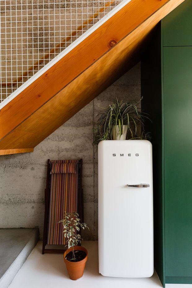 A stylish interior corner beneath a timber staircase or sloped structural element. The scene features a retro white SMEG refrigerator beside dark green built-in cabinetry, a raw textured concrete wall, a striped folding chair, and potted plants. Warm timber and earthy furnishings contrast with the cool concrete and deep green cabinetry.