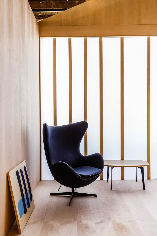 A refined, minimalist interior corner featuring a dark sculptural lounge chair, a small round side table, pale timber flooring and wall panelling, and a full-height translucent window or screen with vertical timber battens. A framed artwork leans against the wall, while exposed dark timber overhead adds contrast.