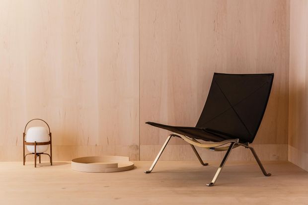 A minimalist interior vignette with pale timber wall and floor panels, featuring a black leather sling chair with a metal frame, a small sculptural side table, and a compact rounded object on a stand. The restrained composition emphasizes natural timber, clean lines, and soft warm lighting.