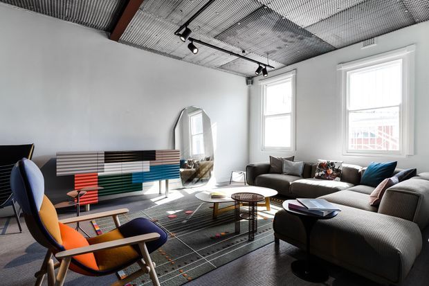 Bright contemporary living room with large windows, a deep grey sectional sofa, colourful mid-century armchairs, patterned rug, low coffee tables, and a colourful horizontal slatted sideboard. The room features an exposed industrial-style perforated metal ceiling with black track lighting and a decorative arched mirror.